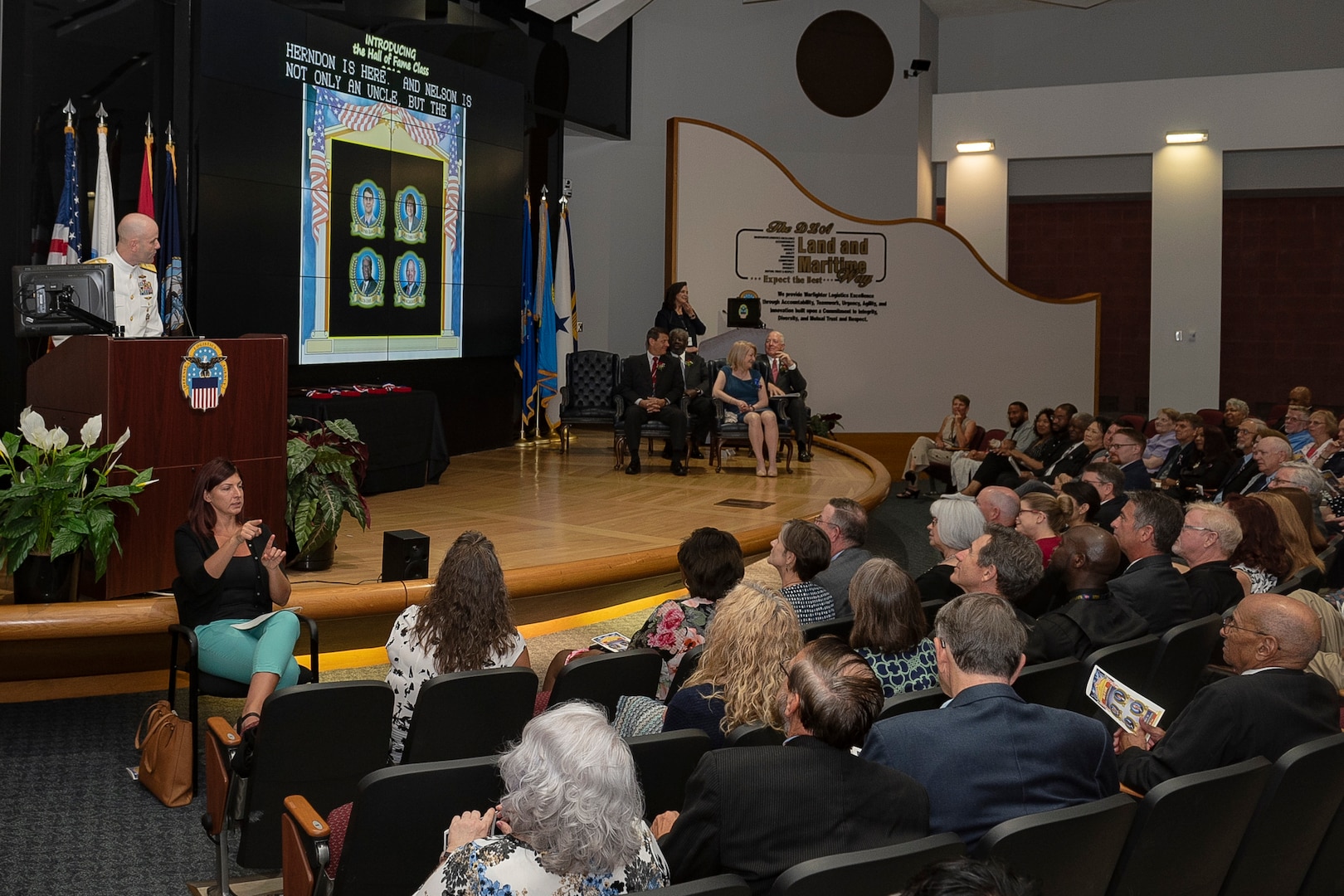 Military man behind a podium on stage speaks to a room full of people