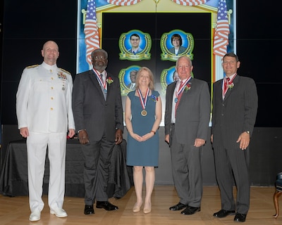 A group of five stand on a stage, one military and four civilians