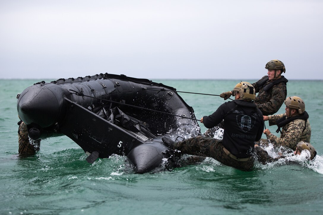 Marines in water use ropes and leverage to pull a small rubber craft closer to them.