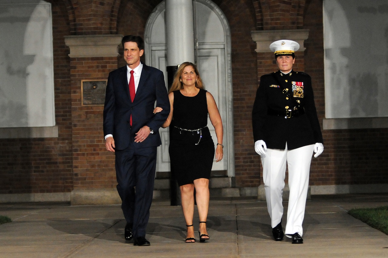 A man and women, with their arms linked, are escorted by a military officer down an outdoor walkway.