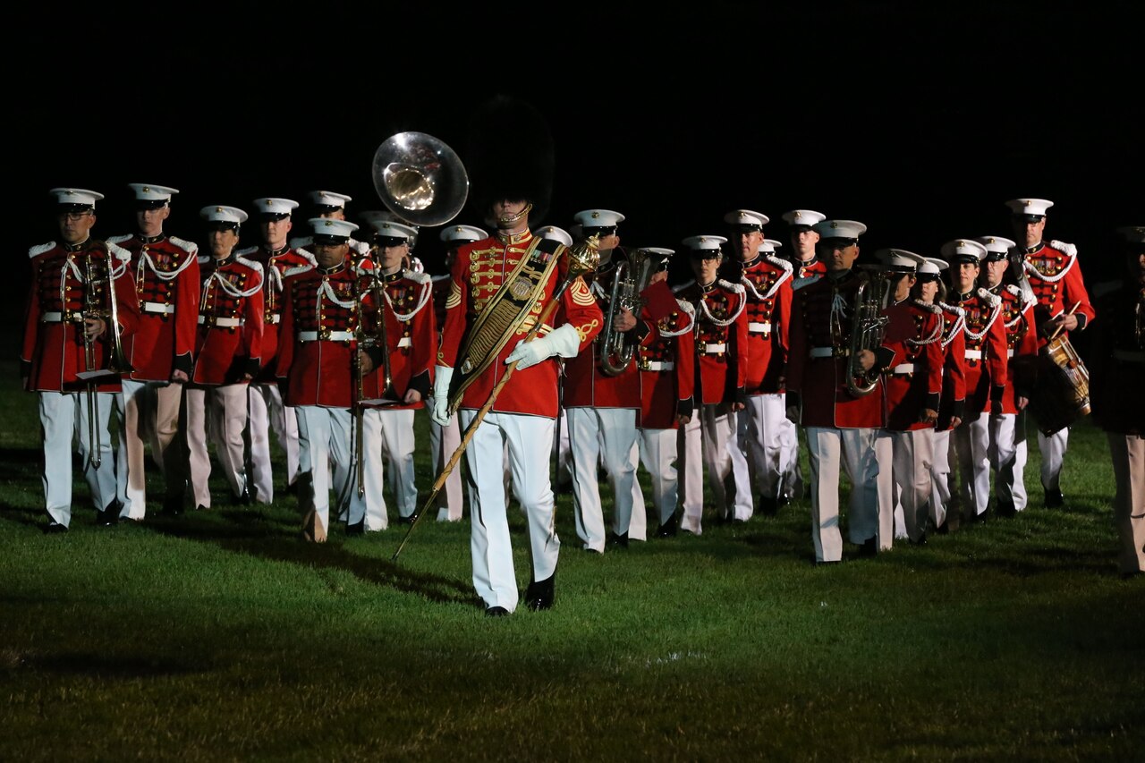 A military band marches, in uniform, across a lawn at night.