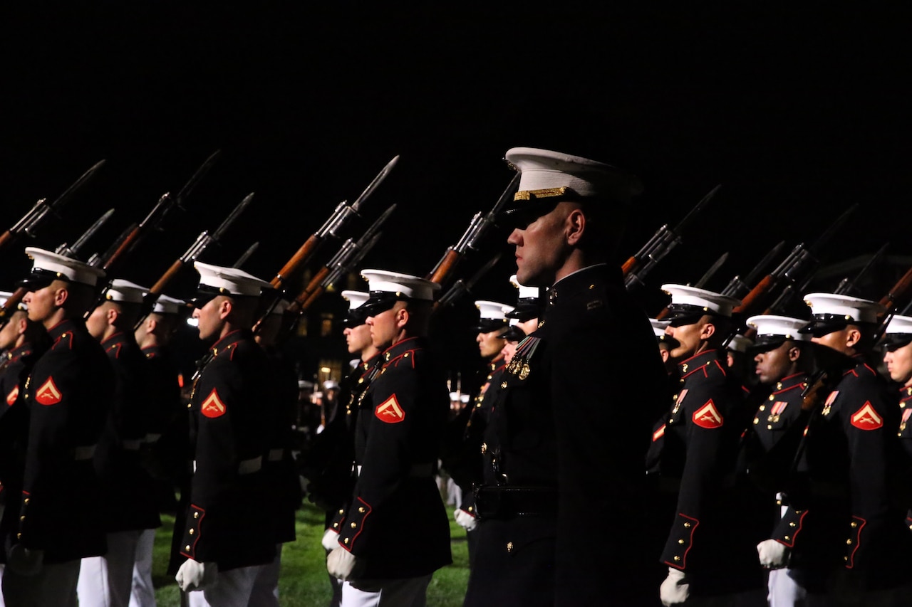 Marines march in formation from right to left wearing white caps and white gloves with rifles slung over their right shoulders.