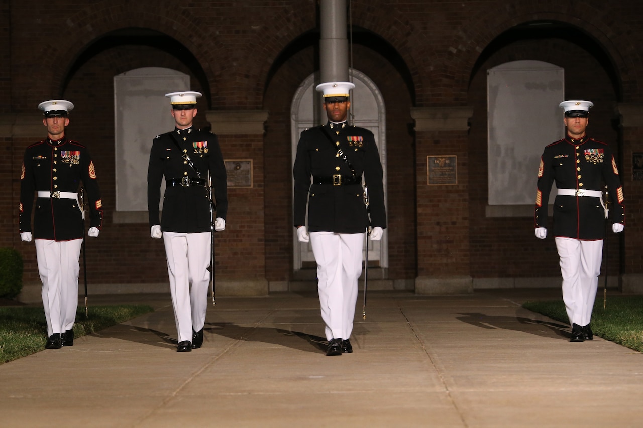 Four service members are lit by a spotlight while lined up on an outdoor walkway in front of a brick building at night.