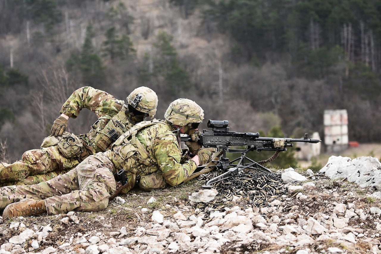 A soldier fires a machine gun from the ground while another reaches for a knapsack.