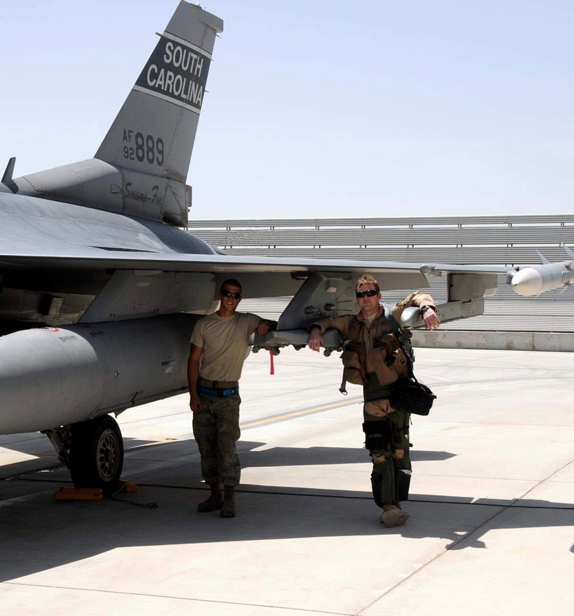 Two Airmen stand  under the wing of an F-16 Fighting Falcon fighter jet.