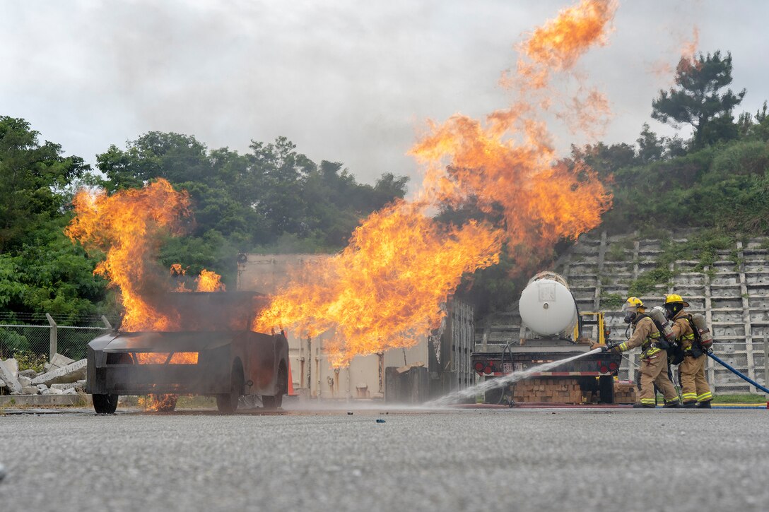 Marines in firefighting gear prepare to point a hose at a fire in a simulated vehicle near a tanker on pavement.