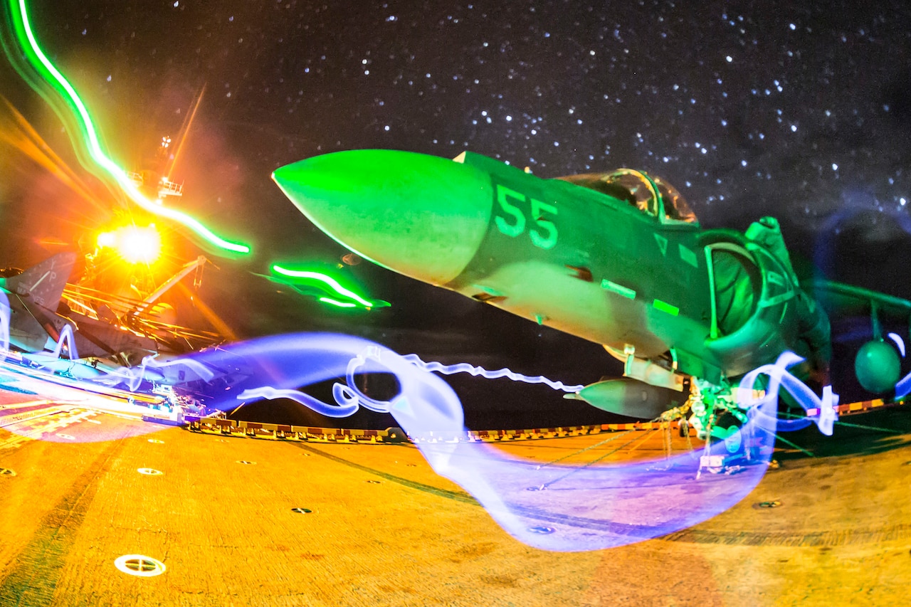 A Harrier jet sits on a ship bathed in colorful light.