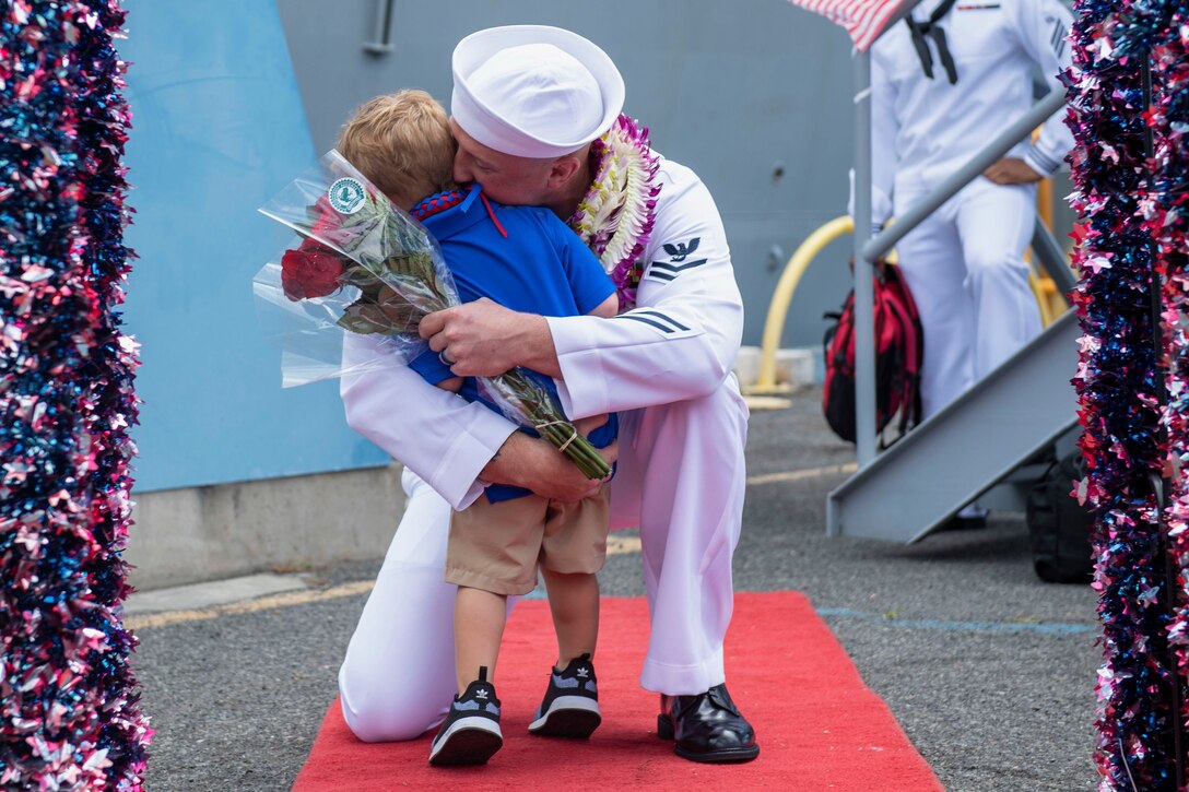 A sailor kneels an a red carpet to hug a child holding flowers.