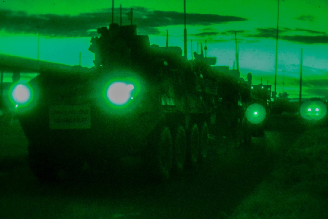 Military vehicles wait in line with their lights shining in a greenish twilight.
