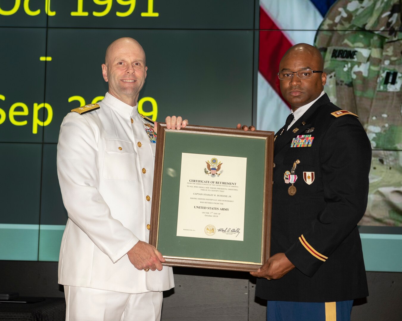 A Navy and Army officer stand on stage holding framed certificate