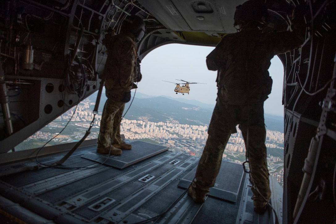 Two soldiers look out the back of a helicopter at another helicopter while flying over a city.