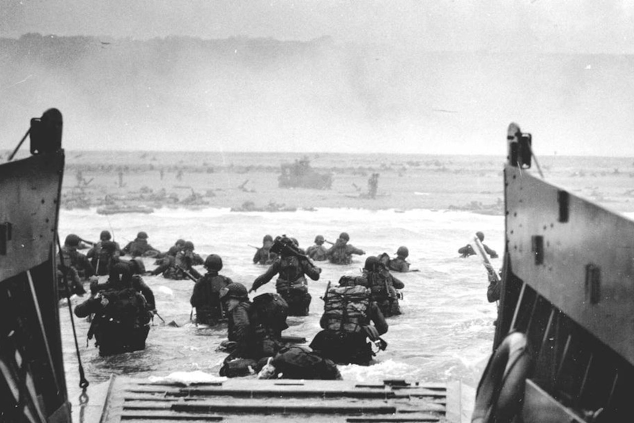 Soldiers wade into waist-deep water toward the shore of a beach from a ship with a ramp in the back.