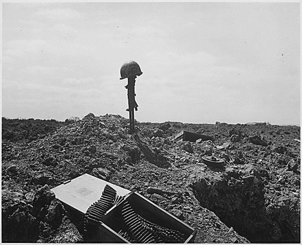 A soldier’s rifle sticks straight up in the sand on a beach with a helmet on it. A box of ammo sits in the foreground.