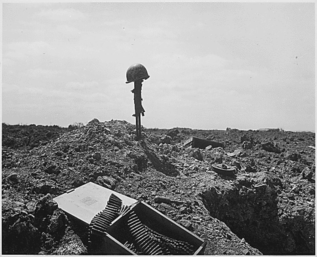 A soldier’s rifle sticks straight up in the sand on a beach with a helmet on it. A box of ammo sits in the foreground.