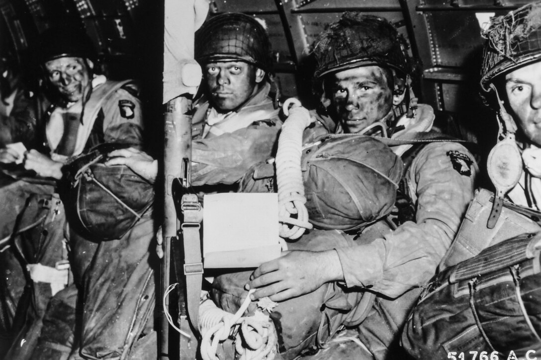 Four paratroopers in face paint sit holding their parachutes in a plane as they prepare to jump on D-Day. One of them holds a note that is his orders from Army Gen. Dwight D. Eisenhower.