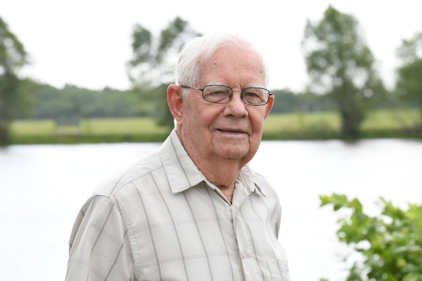 A World War II veteran wearing glasses stands in front of a lake.