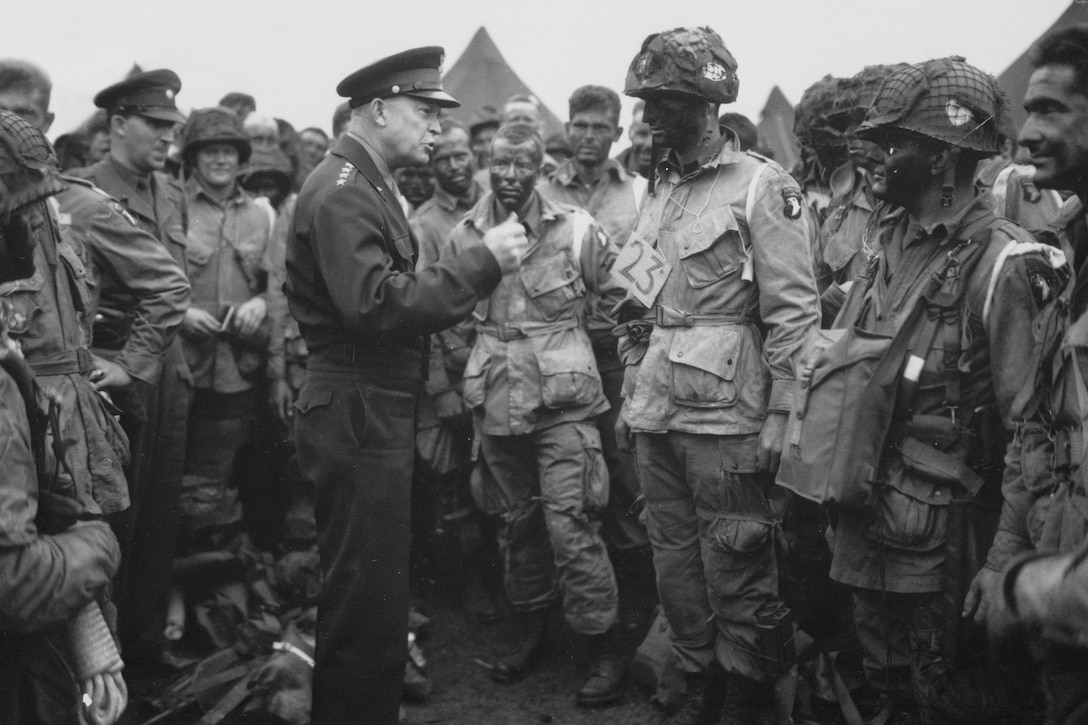Army Gen. Dwight D. Eisenhower talks to soldiers standing around him outside.