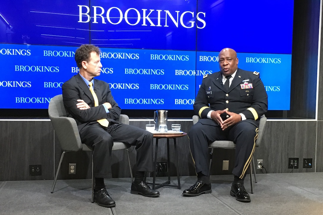 An Army general and a civilian sit on an elevated platform.