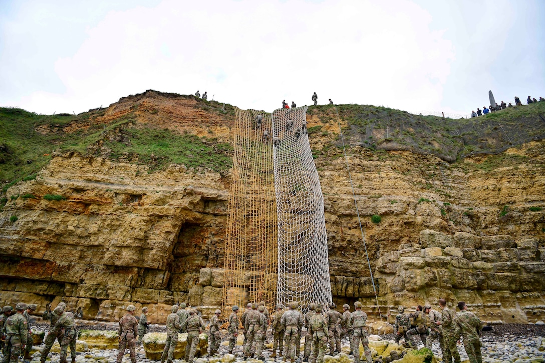 Soldiers climb a cliff using net ladders as others watch from below.