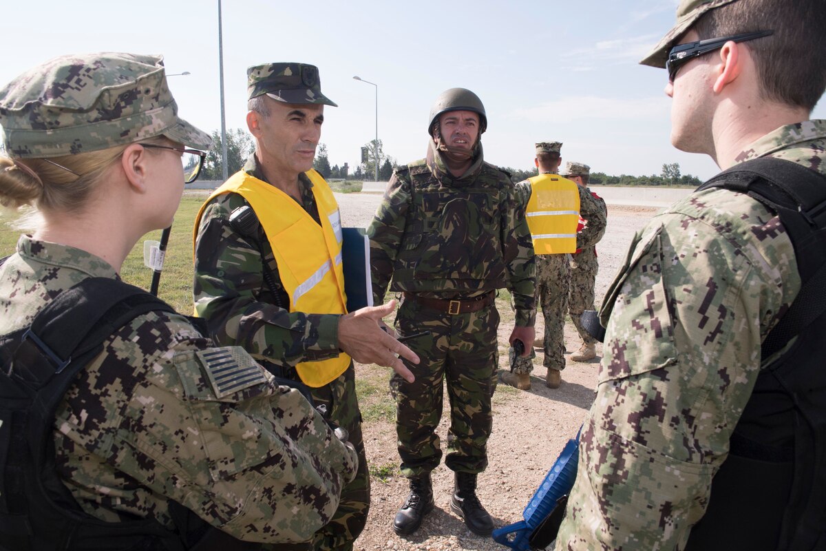 A uniformed service member wearing a yellow vest addresses service members wearing U.S. military uniforms.