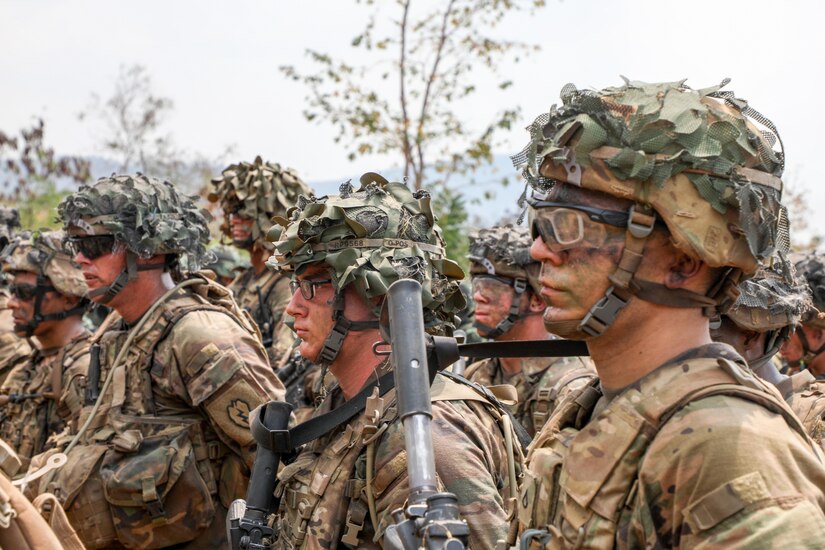 Military personnel in camouflage uniforms stand outdoors in formation.