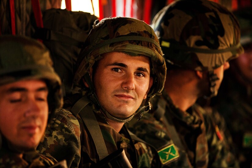 Military personnel in camouflage uniforms and combat helmets sit side by side in a helicopter.