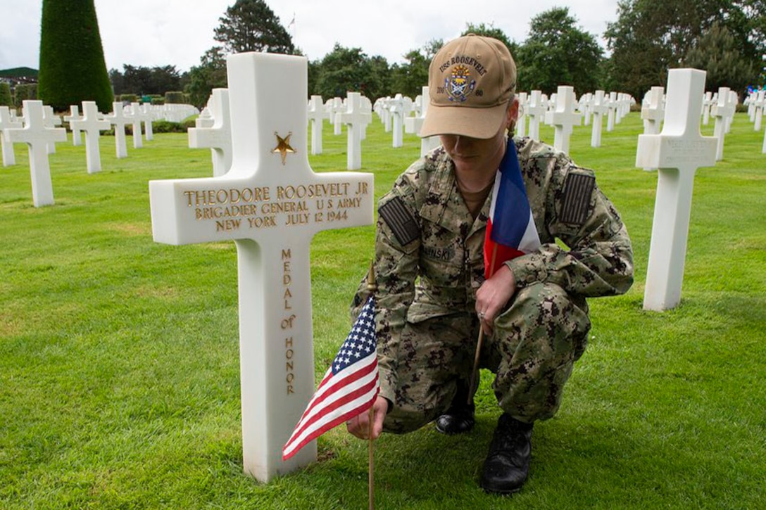 A sailor places U.S. and French flags in front of headstones in a cemetery.