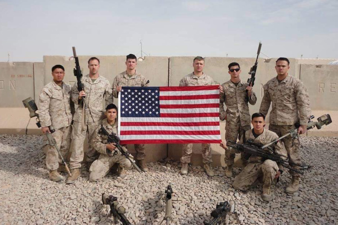 Eight Marines pose with an American flag in front of a cement barricade in Afghanistan.