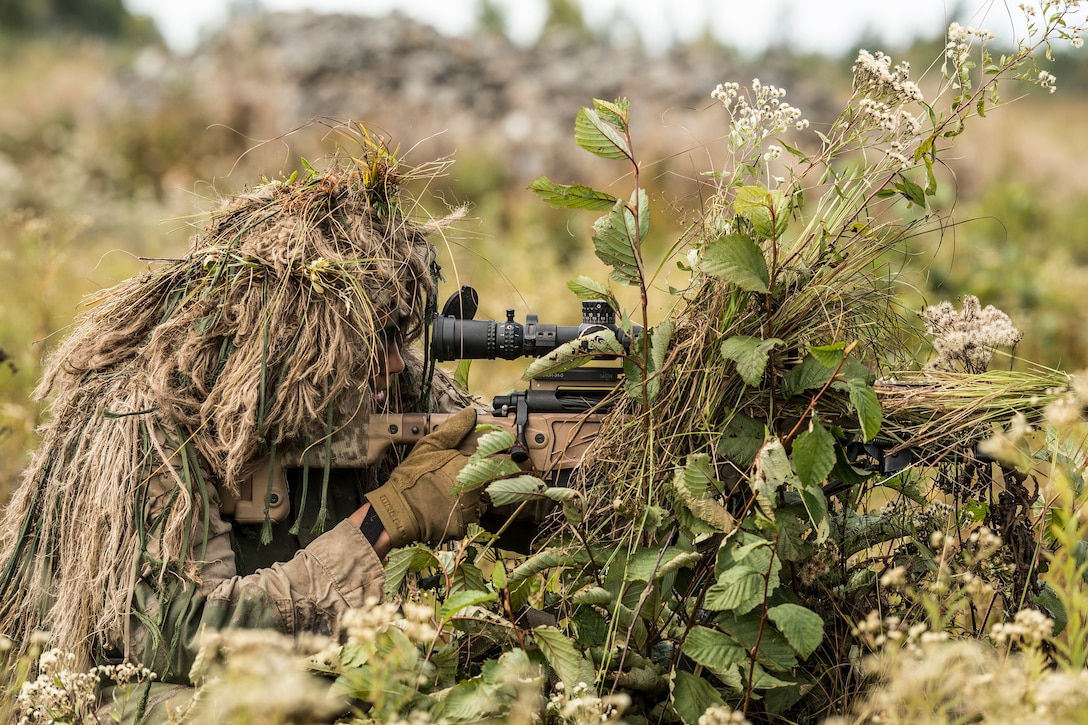 A Marine, camouflaged by a ghillie suit  looks through the sight of his sniper rifle.