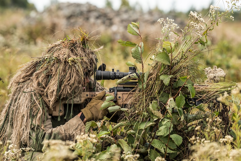 A Marine, camouflaged by a ghillie suit  looks through the sight of his sniper rifle.