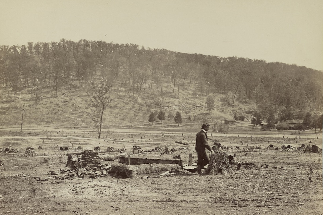 A man walks in a field in front of a ridge covered in trees.