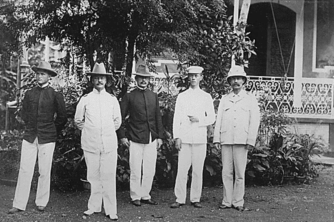 Five men pose for a photo in front of a building, trees and a shrub.