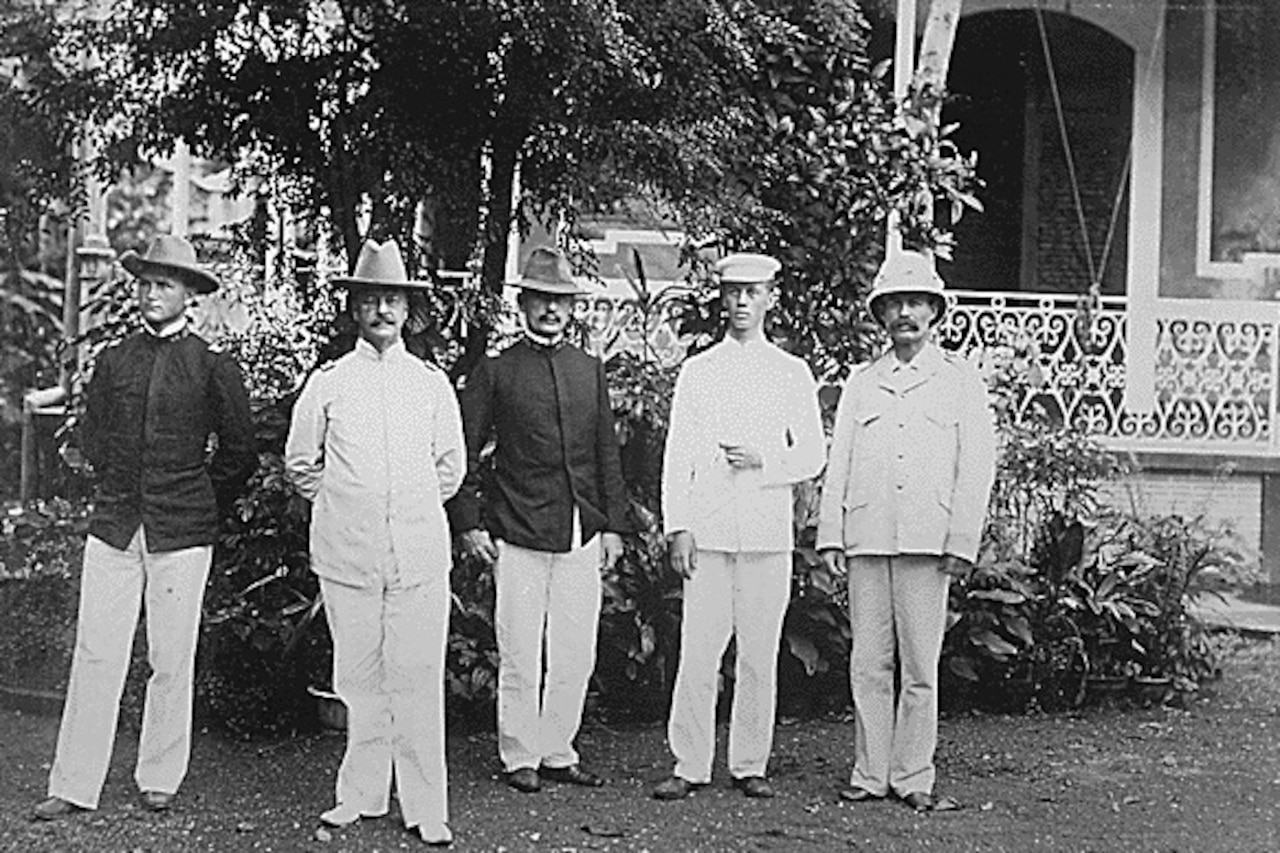 Five men pose for a photo in front of a building, trees and a shrub.