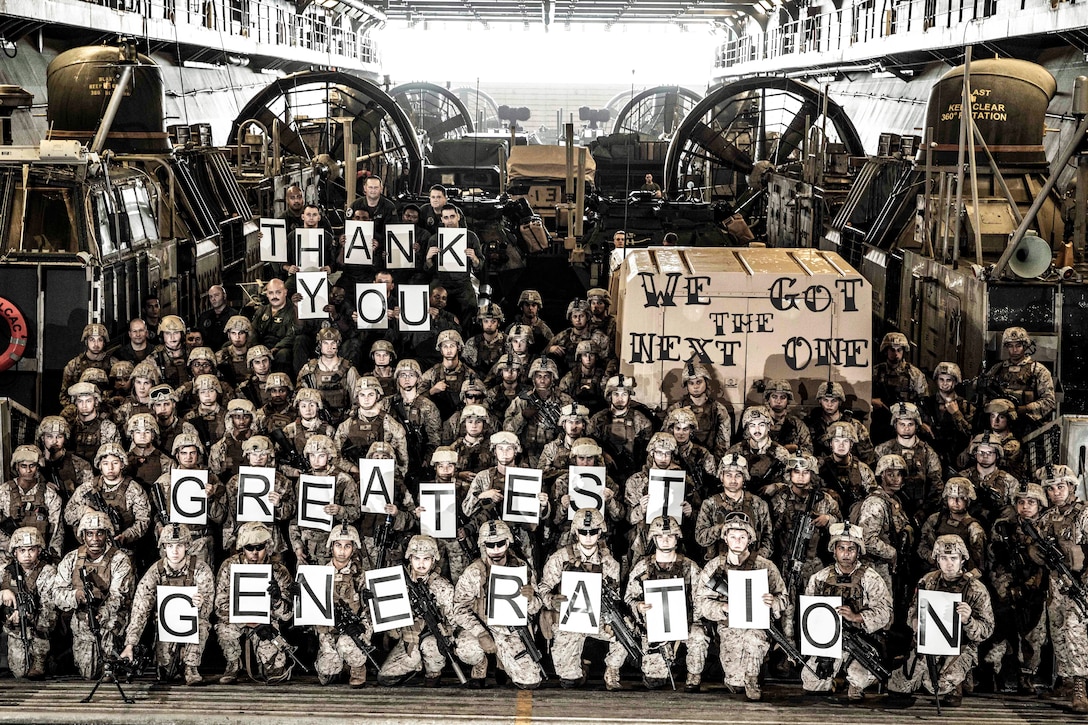 A large group of sailors pose for a photo while some hold up signs.