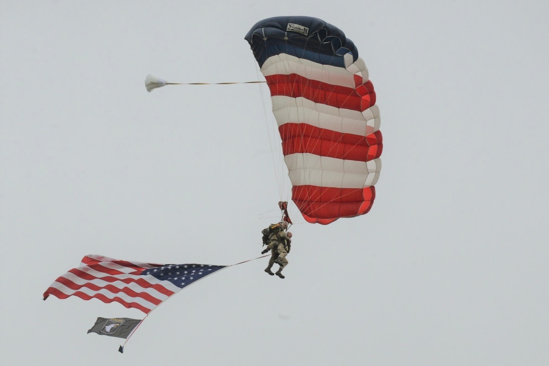 Tandem parachutists attached to an American flag parachute descend in the sky with an American flag trailing them.