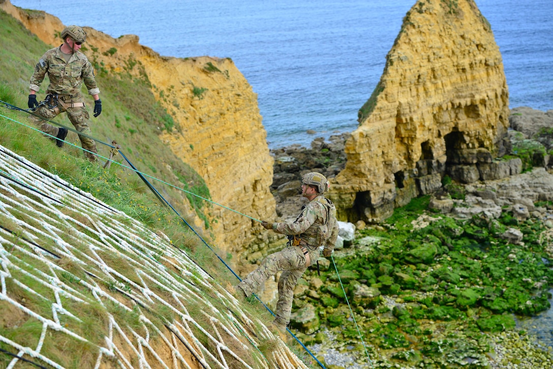 A soldier uses a rope to climb a steep bluff overlooking the ocean as another soldier watches.