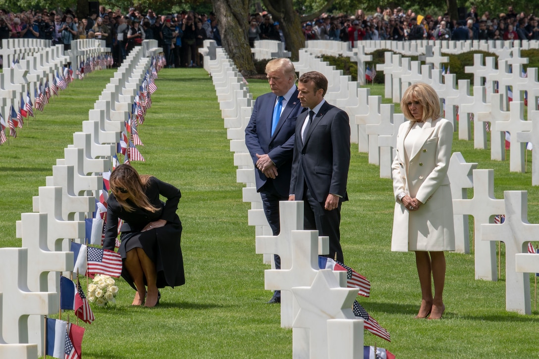 A woman crouches to lay white flowers at a graveside as heads of state and a distant crowd look on.