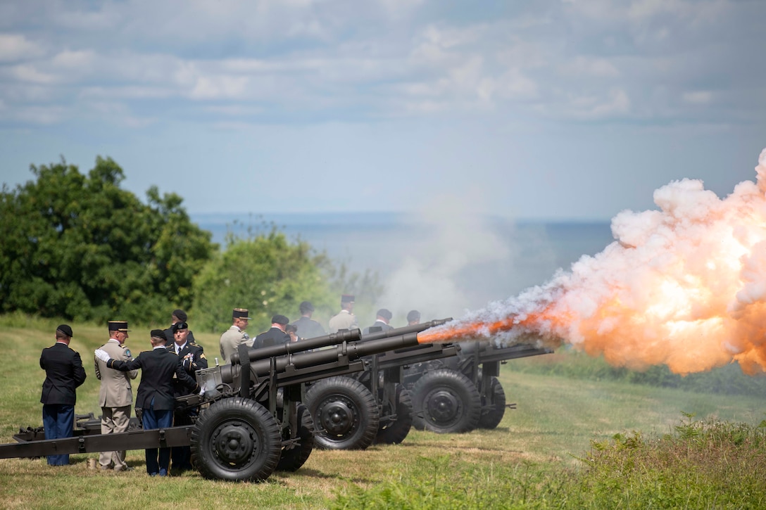 Soldiers fire cannons in a field overlooking the ocean.