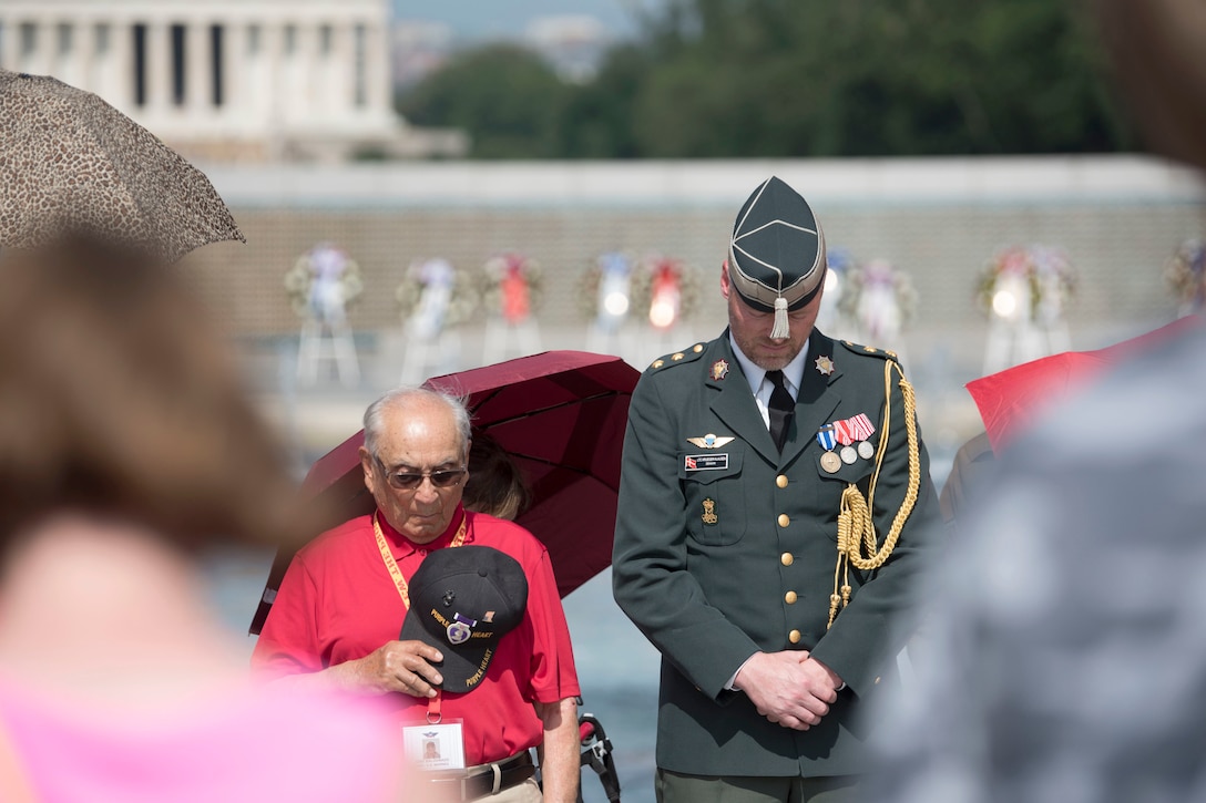 A service member and veteran bow their heads at a memorial.