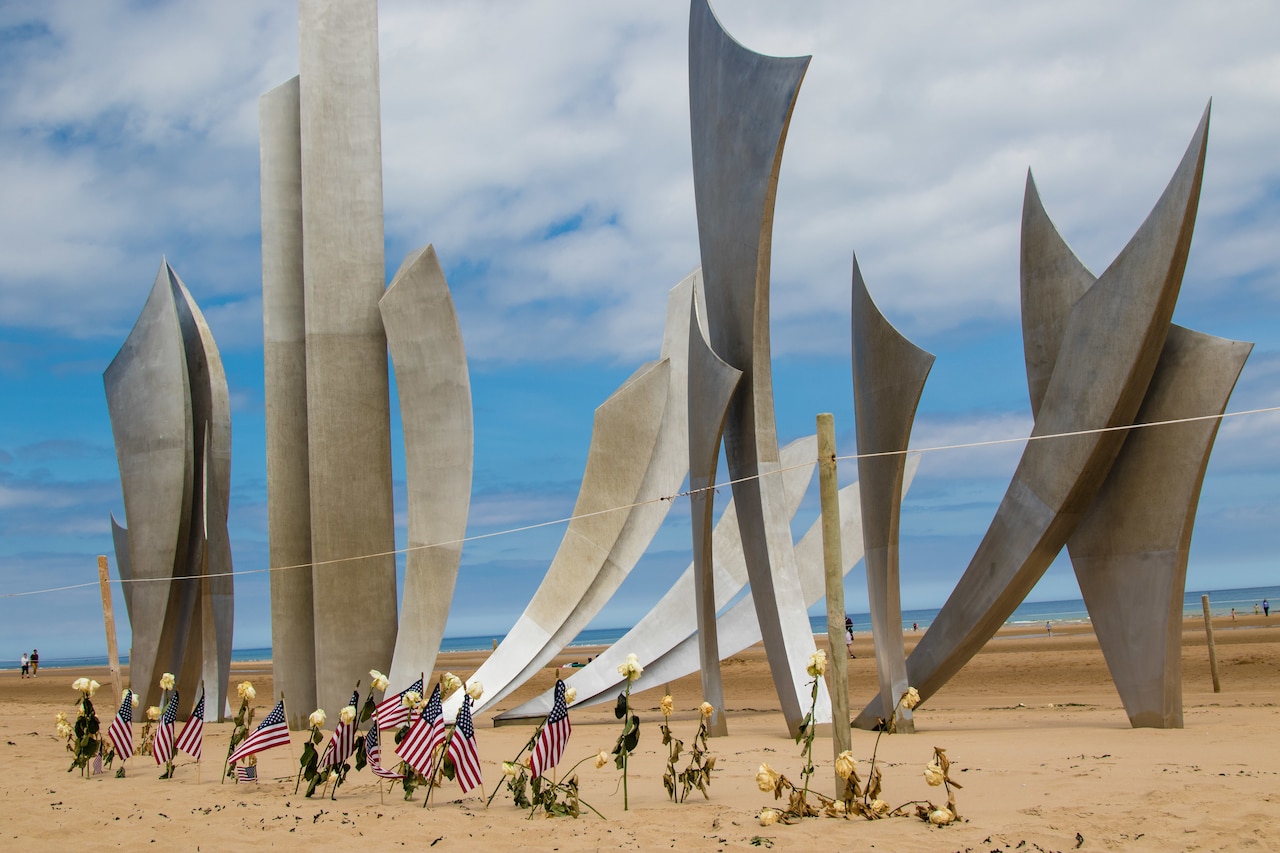 A sculptured memorial on the beach with flowers and American flags planted in the sand.