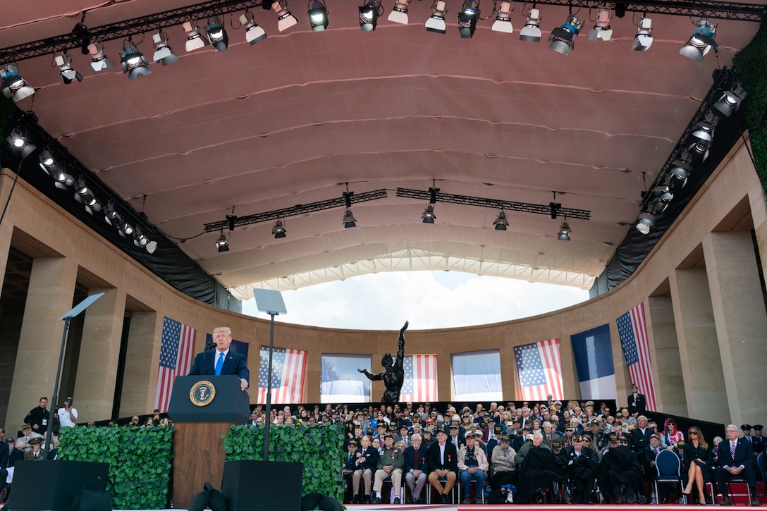 President Donald J. Trump speaks from a podium in a large hall surrounded by WWII veterans and American flags.