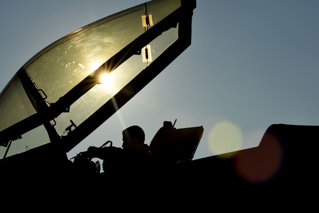 A pilot sits in an open cockpit holding a helmet as the sun reflects off glass.