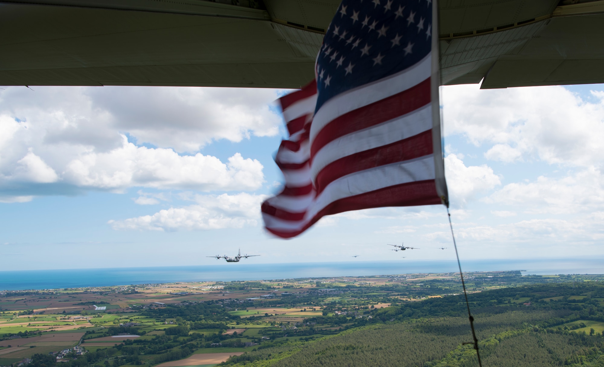 The American flag, secured to the open ramp of the lead 37th Airlift Squadron C-130J Super Hercules, waves in the wind, as seven C-130J Super Hercules tail behind as part of an eight-ship formation in Normandy, France, June 6, 2019. The eight-ship formation flyover was part of the commemoration events in honor of the 75th anniversary of D-Day. In this formation, four C-130J Super Hercules belong to the 37th AS out of Ramstein Air Base, two belonging to the 61st and 62nd AS out of Little Rock Air Force Base, and two belonging to the 39th and 40th AS out of Dyess Air Force Base. (U.S. Air Force photo by Senior Airman Kristof J. Rixmann)