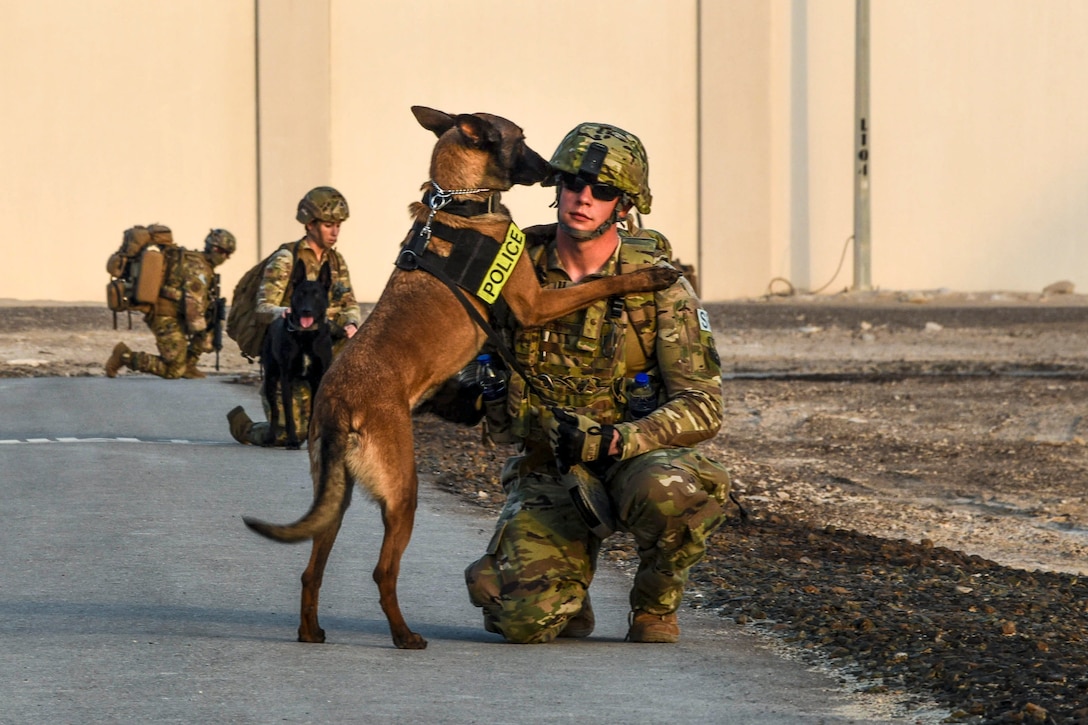 A military dog places his front paws on a kneeling airman on a road with other kneeling airmen behind him.