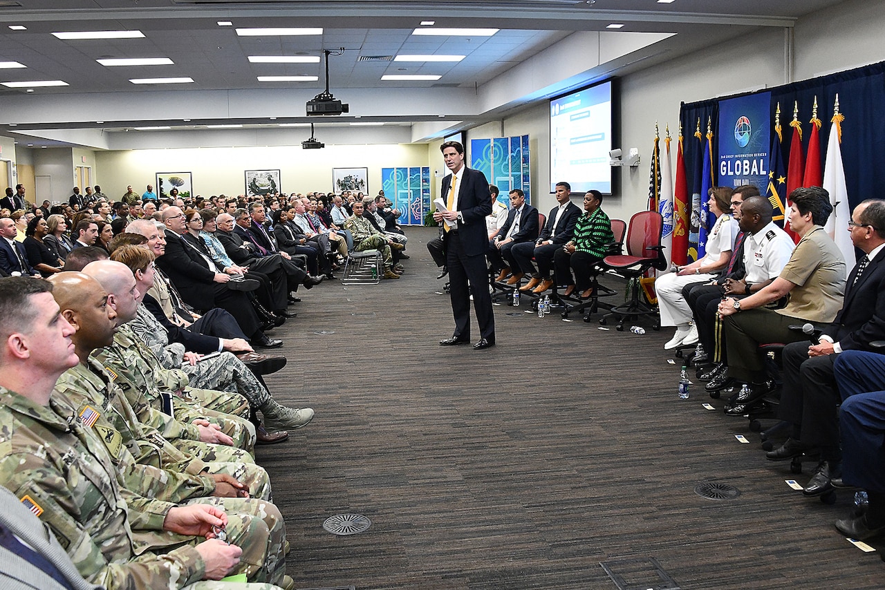 Man speaks to a conference room full of military and civilian personnel.