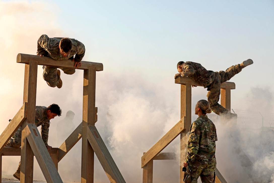 A soldier watches three other soldiers climb over an obstacle with smoke in the background.