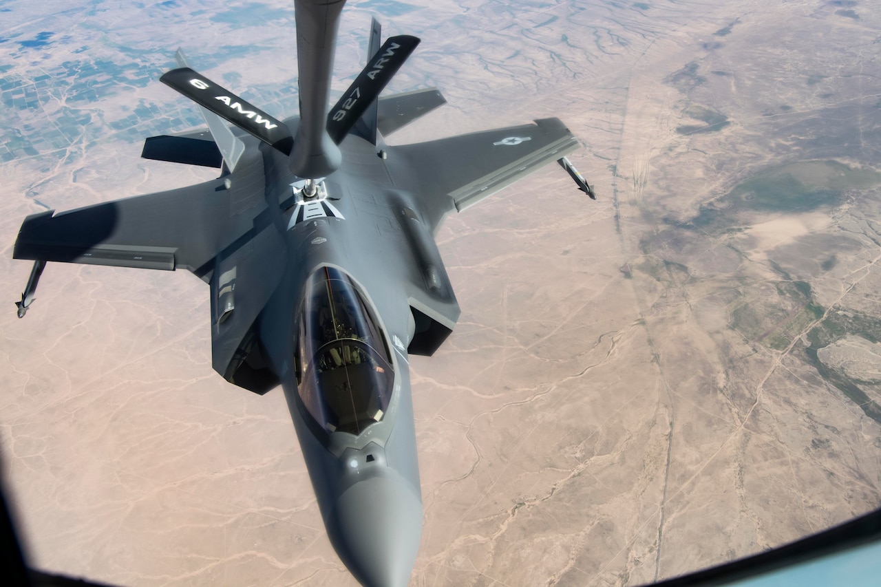 A fighter aircraft is refueled in mid-air over a desert-like landscape.