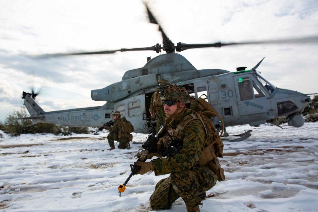 A service member with a rifle squats in the snow as other personnel exit a helicopter.