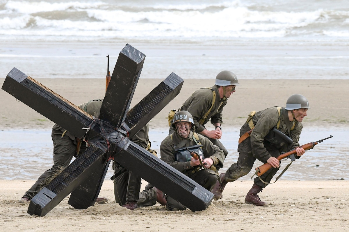 A group of sailors in 1940s uniforms run on a beach near a big metal obstacle.