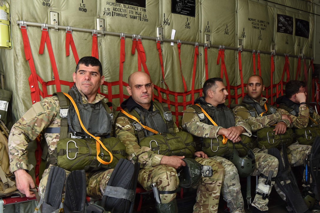 Service members with parachute bags on their laps sit against a wall onboard an aircraft.